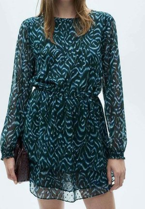 Femme portant une robe en voile bleu sarcelle foncé avec des motifs abstraits bleus et blancs, manches longues, taille cintrée, et tenant une pochette texturée marron foncé.