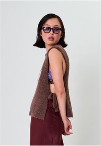 Pruun kootud varrukateta vest pehme tekstuuriga, õrna avatud selja ja paelaga detailiga. Kandmisega paaritatud lilla topi ja burgundi seelikuga.