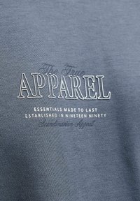 Μπλε ύφασμα με λευκό κείμενο που γράφει "APPAREL essentials made to last established in nineteen ninety" με έντονη και μικρή γραφή.