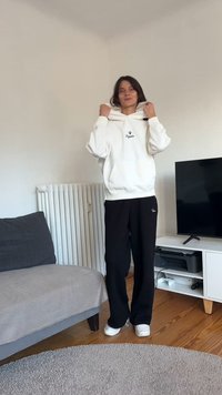 Camisola com capuz branca com logótipo, combinada com calças pretas caneladas. Tênis brancos. A sala inclui um sofá cinzento, radiador e móvel para TV. Piso de madeira.