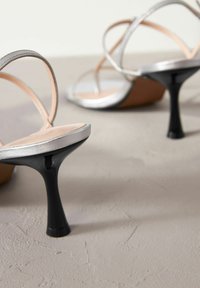 Zilveren sandalen met hakken en dunne bandjes, met een puntige zwarte hak en een glad metallic uiterlijk. Zacht beige voetbed completeert het ontwerp.
