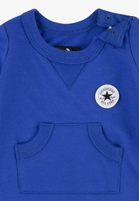 Sweatshirt azul com gola redonda canelada, detalhe em botão, bolso frontal e um emblema circular com o logótipo "Converse All Star". Tecido macio.