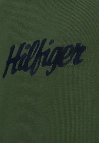 Zielona dzianina z charakterystycznym granatowym logo „Hilfiger”, o teksturowanym splocie i prostym wzorze.