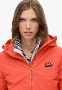 Chaqueta roja impermeable con cierre frontal, capucha ajustable y parche de logo. Superpuesta sobre una camisa de rayas con cuello. Textura suave.