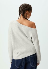 Grauer schulterfreier Pullover mit lockerer Passform, geripptem Saum und weicher Textur. Kombiniert mit dunkelblauen Jeans mit Gesäßtaschen.