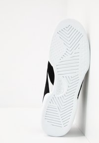 Baskets noires avec une semelle en caoutchouc texturée blanche, une tige noire lisse et un détail de logo blanc sur le côté.