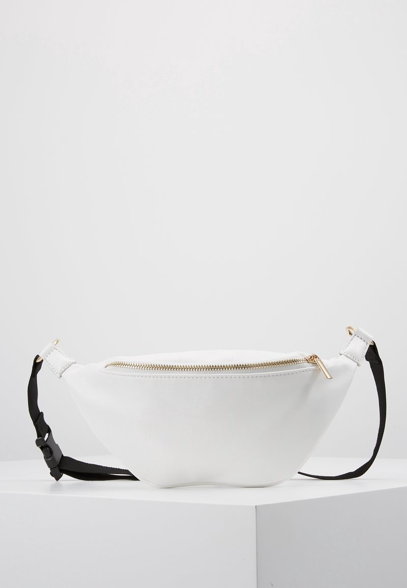 Sac banane en cuir blanc avec une fermeture éclair dorée et une sangle noire ajustable, posé sur une surface blanche sur fond gris clair.