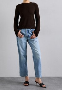 Braun gestrickter Langarm-Oberteil mit gewelltem Ausschnitt und Bündchen, kombiniert mit hellblauen geraden Jeans. Schwarze hochhackige Mules runden das Outfit ab.