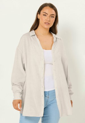 Vrouw die een oversized lichtbeige overhemd met knopen draagt, een witte tanktop en lichtblauwe spijkerbroek, staande tegen een effen achtergrond.