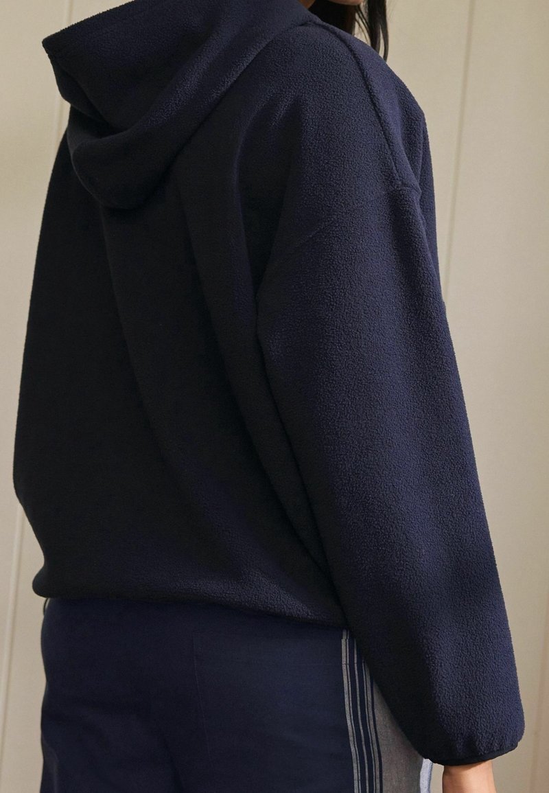 Osoba ubrana w granatowy polarowy hoodie i pasujące spodnie z jasnoszarymi paskami po bokach, stojąca na tle jasnej, jednolitej ściany.
