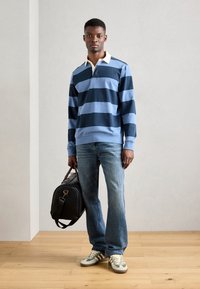 Polo à rayures à manches longues dans des tons de bleu, avec un col crème ; associé à un jean bleu délavé et des baskets avec des accents verts ; tenant un sac noir.
