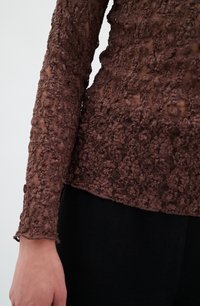 Maglietta lunga a maniche con texture marrone, decorata con un motivo floreale e dettagli trasparenti, abbinata a un capo nero sottostante. Aspetto morbido e stratificato.