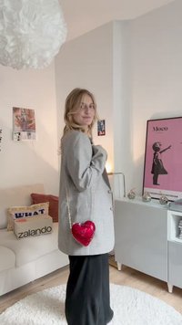 Grijze geruite blazer met een glanzende rode hartvormige tas aan een gouden ketting. Zwarte broek en een lichte achtergrond met decoratieve elementen.