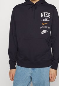 Marinblå hoodie med framficka, prydd med flera Nike-logotyper i vitt, orange och svart. Inkluderar en huva med dragsnodd och ribbade ärmslut.