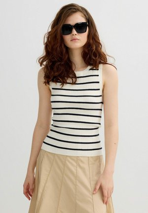 Mujer con cabello castaño ondulado, usando gafas de sol negras, top blanco sin mangas con rayas horizontales negras y falda beige, sobre un fondo liso.