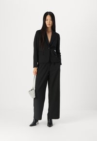 Completo nero su misura con giacca aderente e pantaloni a gamba larga, accessoriato con una borsa argentata a trama e stivaletti con punta.