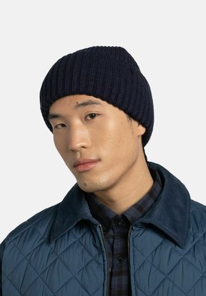 Navy ribbed knit beanie with a snug fit, paired with a blue quilted jacket and plaid shirt. Focus on texture and color contrast.