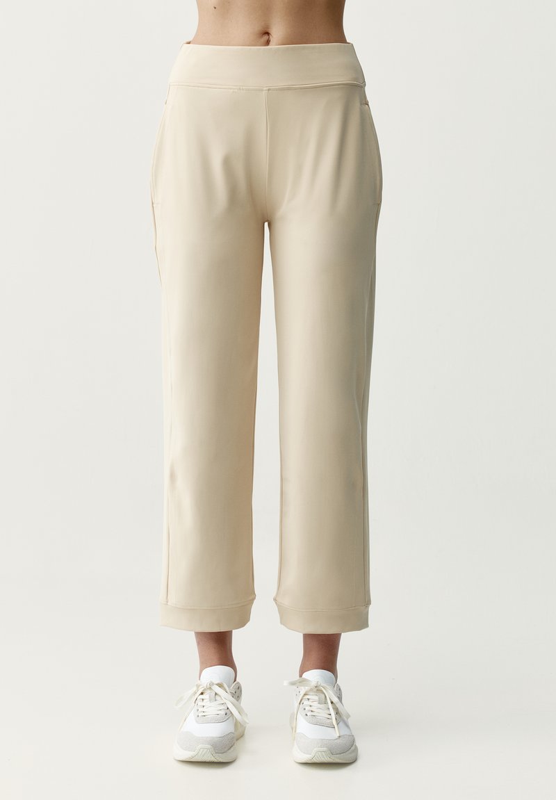 Beige Hosen mit weitem Bein und umgeschlagenen Bündchen, getragen von einer stehenden Person, kombiniert mit weißen und grauen Schnürschuhen vor einfarbigem Hintergrund.