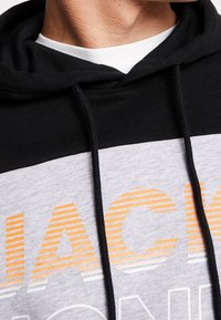 Sudadera con capucha de sección superior negra, cuerpo gris y texto gráfico de rayas naranjas. Cuenta con cordones y una textura suave.