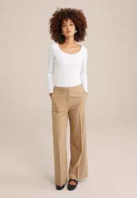 Top de manga larga blanco acanalado combinado con pantalones anchos beige. La modelo está de pie con las manos en los bolsillos, usando zapatos negros con tachuelas.