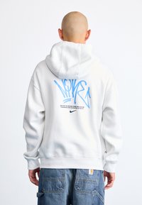 Pessoa com a cabeça raspada vestindo um sweatshirt branco da Nike com texto em estilo graffiti azul e logotipo preto nas costas, combinado com calças de ganga azuis.