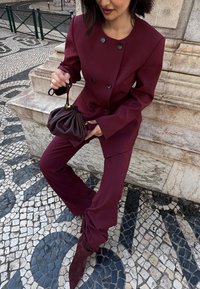 Burgundijas uzvalks ar divrindains dizainu un lieliem pogām, kā arī brūna plīša soma un augstpapēžu zābaki, izvietoti uz rakstainām flīzēm.