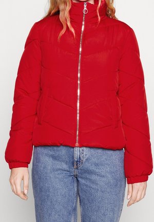 Femme portant une doudoune matelassée rouge avec une fermeture éclair argentée à l'avant et un jean bleu, debout devant un fond uni.