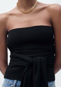 Top nero a coste senza spalline con dettaglio annodato in vita. Abbinato a pantaloncini di jeans. Presenta una superficie liscia e un design aderente.