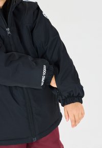 Schwarze wasserdichte Jacke mit elastischen Bündchen, ausgestattet mit einer Reißverschlusstasche und einem "WPRO 10000" Logo auf dem linken Arm.