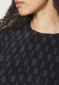 Gros plan sur le bas du visage et le cou d’une femme aux cheveux bouclés, portant un haut noir à motifs de rectangles gris foncé.