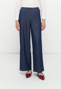 Pantaloni in denim a gamba larga di colore blu scuro, con vita regolabile tramite cordoncino, cuciture a contrasto e una texture liscia. Abbinati a scarpe rosse.