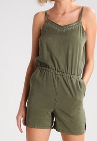 Olivově zelený kombinéza s nastavitelnými tenkými ramínky, elastickým pasem a dekorativním prošíváním podél výstřihu. Lehká a pohodlná látka.