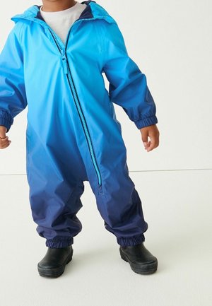 Combinaison imperméable bleue avec un dégradé du bleu clair au bleu foncé, dotée d'une fermeture éclair à l'avant et de poignets élastiques, portée avec des bottes noires.