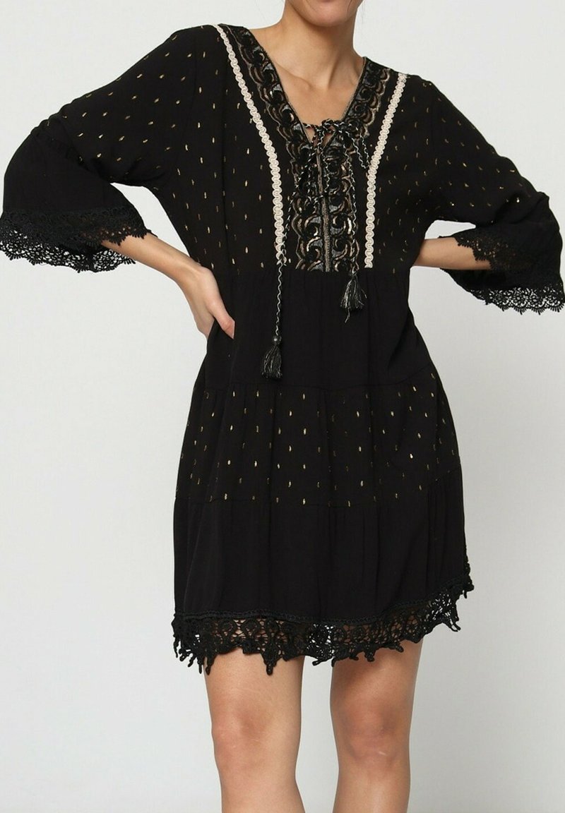 Robe noire avec bordure en dentelle, détail de décolleté complexe et accents dorés. Design fluide avec des manches trois-quarts.