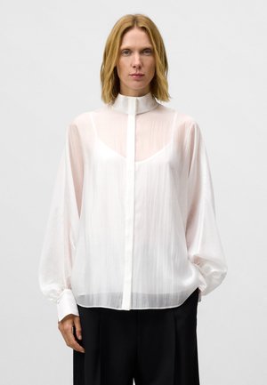 Blusa bianca e trasparente con colletto alto, chiusura a bottoni e maniche lunghe e svolazzanti. Tessuto morbido e fluido con una leggera lucentezza.