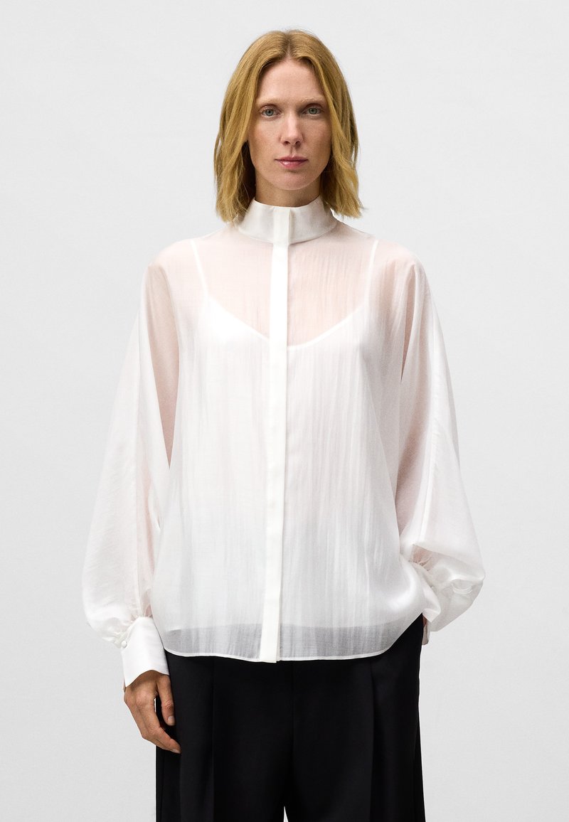 Witte, doorschijnende blouse met een hoge kraag, knoopsluiting aan de voorkant en lange, luchtige mouwen. Zacht, vloeiend materiaal met een lichte glans.