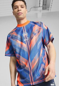 Sarkans ar īsām piedurknēm, zilā un oranžā krāsā, ar ģeometriskiem rakstiem, apaļo kakla izgriezumu un Puma logotipu uz krūšu daļas, izgatavots no sintētiska materiāla.