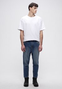 White cotton t-shirt with short sleeves, paired with light blue denim jeans and black lace-up boots, standing against a neutral background.