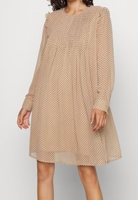 Beiges Kleid mit Punkten aus leichtem Stoff, mit langen Ärmeln, Rüschendetails an den Schultern und einer lockeren, fließenden Silhouette.