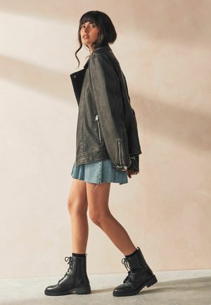Chaqueta de cuero negra con bolsillos con cremallera, combinada con una falda de mezclilla azul clara. La modelo lleva botas de combate negras con cordones.