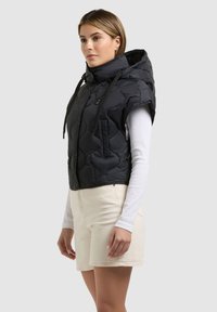 Kvinde iført sort quiltet vest uden ærmer med hætte over hvid bluse med lange ærmer og cremefarvede shorts, stående mod en ensfarvet baggrund.