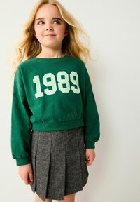 Grüner Pullover mit der Aufschrift "1989" in weißen Buchstaben, kombiniert mit einem grauen Faltenrock im Fischgrätenmuster. Weicher Stoff, lässige Passform.