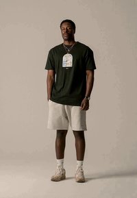 Juoda trumpomis rankovėmis marškinėliai su grafiniu dizainu, derinami su šviesiai pilkomis šortais ir smėlio spalvos sportbačiais, stovintys neutralioje aplinkoje.