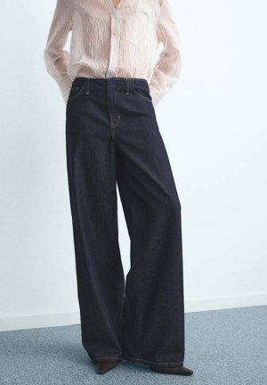 Persona lleva unos pantalones de mezclilla oscuros de pierna ancha y cintura alta, combinados con una camisa de botones de rayas claras y zapatos de punta.