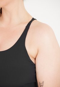 Zwarte sporttop met dunne banden, een gladde textuur, strakke pasvorm en een zichtbare tatoeage op de arm onder de halslijn.