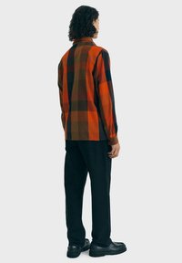 Personne avec des cheveux bouclés portant une chemise à carreaux orange, marron et noir, un pantalon noir et des chaussures noires, tournant le dos face à un fond uni.