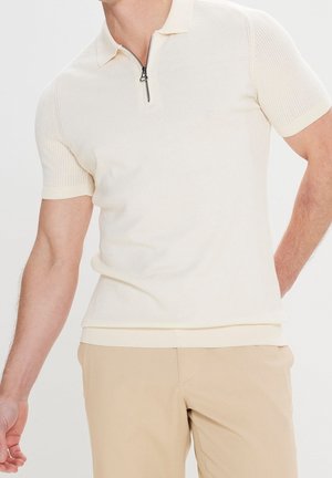 Mann trägt ein cremefarbenes, kurzärmeliges Strick-Poloshirt mit schwarzem Reißverschluss, kombiniert mit hellbeigen Hosen.