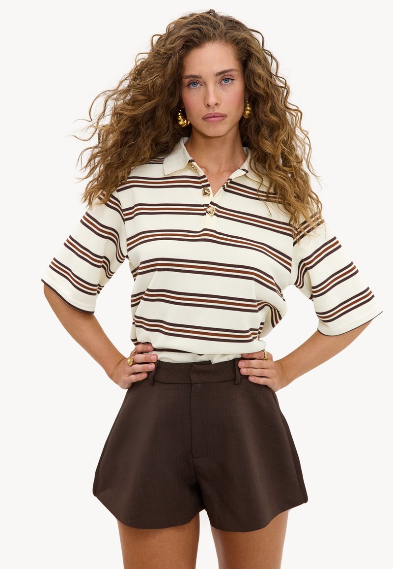 Femme aux cheveux bouclés portant un polo blanc à rayures marron et un short marron foncé taille haute, posant avec les mains sur les hanches.
