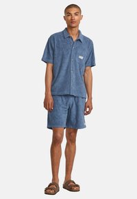 Blue floral-patterned short-sleeve shirt and matching shorts, made of textured fabric, paired with brown sandals. Small logo detail visible.