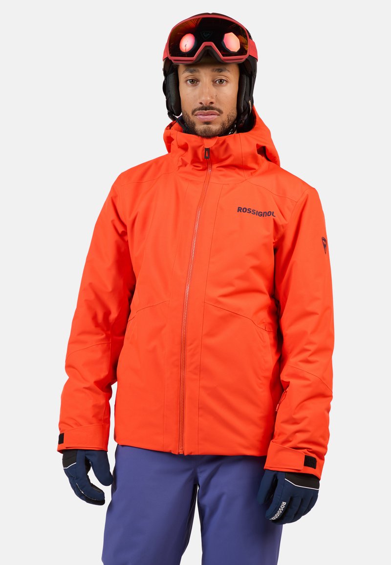 Felgekleurde oranje geïsoleerde skijack met een fitted capuchon, ritssluiting en zwarte accenten, gecombineerd met blauwe skibroeken en zwarte handschoenen.